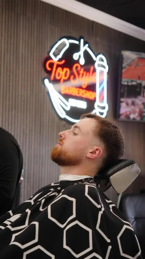 Client leaning back after a fade and beard trim, the red Top Style neon sign behind him